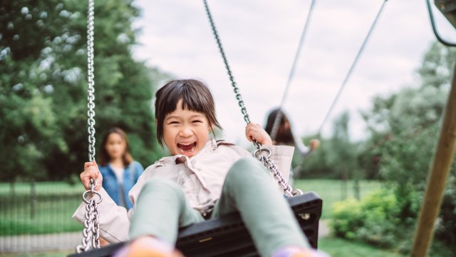 Article thumbnail: Lovely little girl smiling at the camera while playing on a swing set in playground joyfully.